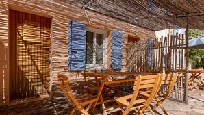 Un espace extérieur aménagé avec des meubles en bois, comprenant des tables et des chaises, un toit de chaume et une fenêtre ornée de volets bleus.