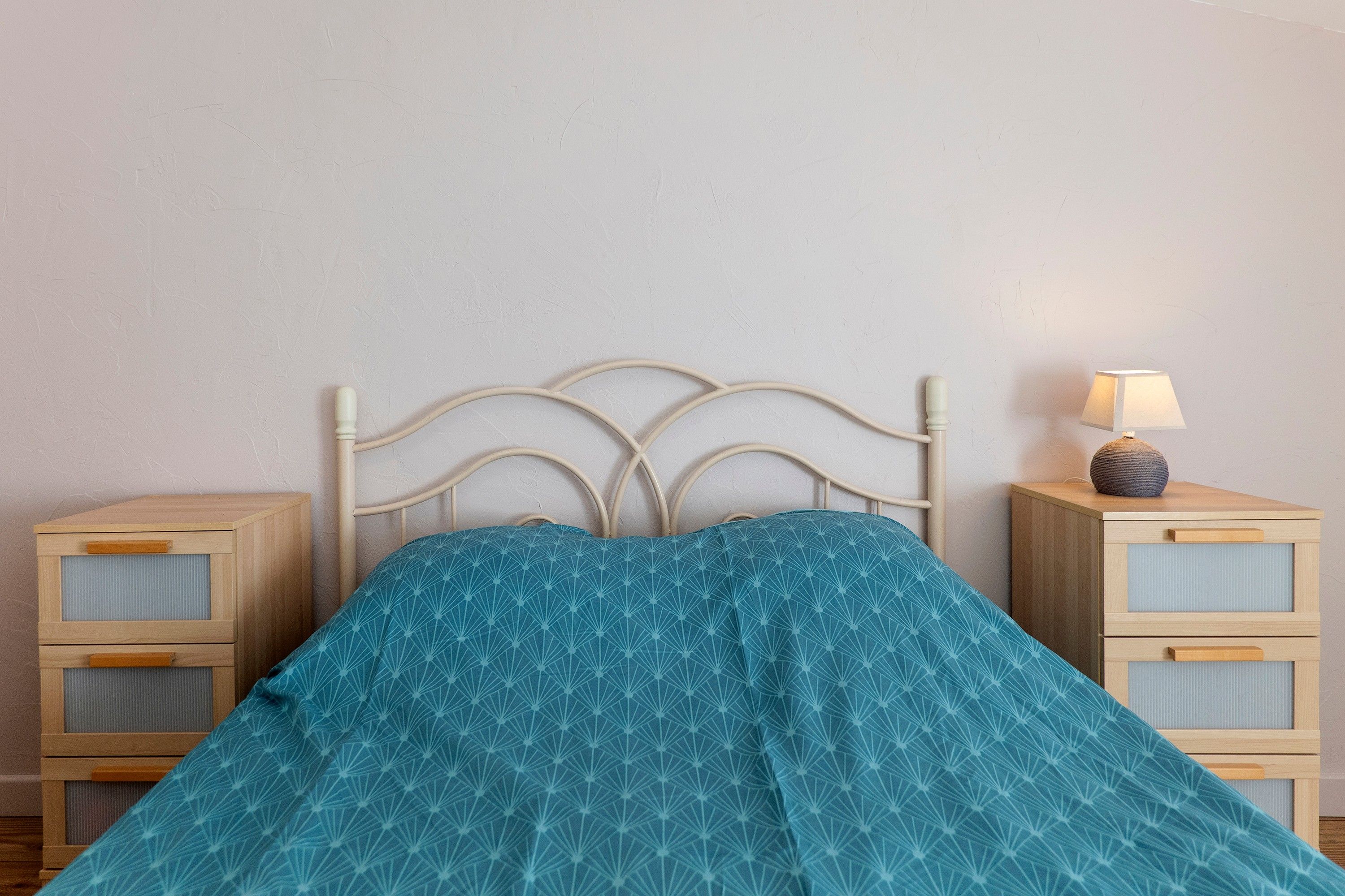 Uma cama com cabeceira, mesa de cabeceira com luminária e conjunto de gavetas.