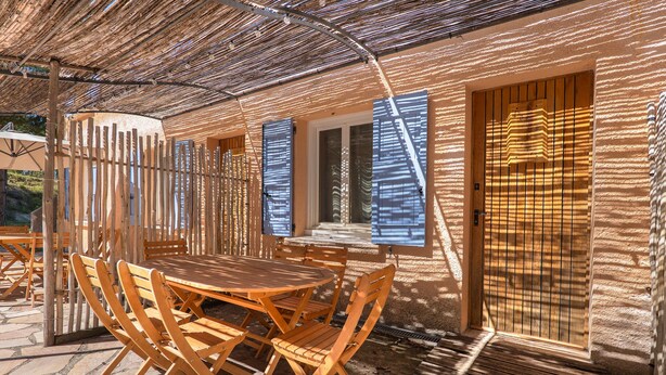 Un espace de repas extérieur en bois, avec une table et des chaises, une pergola au-dessus et une fenêtre avec des stores.