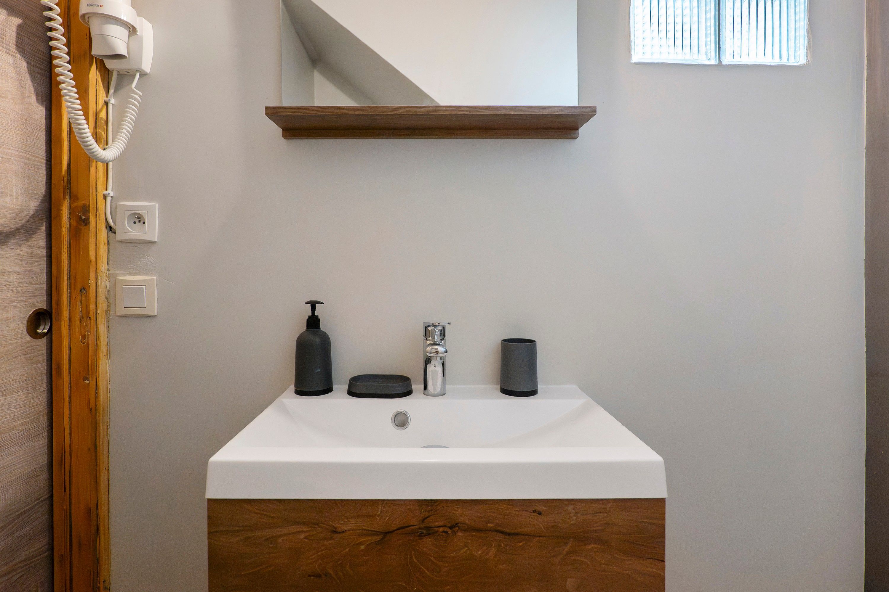 Un baño con un lavabo blanco, un mueble de tocador de madera, un espejo, un secador de pelo y una ventana con persianas.