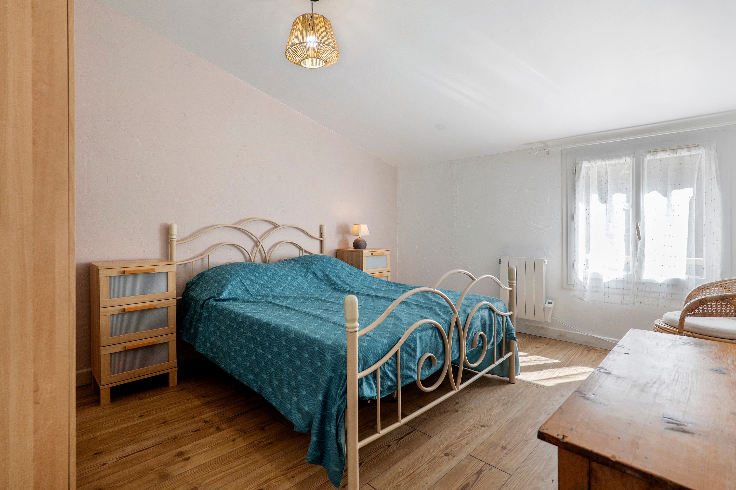 Ein Schlafzimmer mit einem Bett, Nachttischen, einer Lampe, einem Fenster mit Vorhängen und Holzboden.