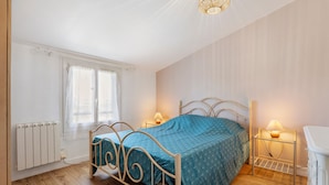 Une chambre à coucher avec un lit en métal, une couvre-lit bleue, deux lampes de chevet, un radiateur et une fenêtre avec des rideaux.