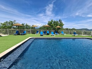 En swimmingpool med blå liggestole og parasoller, omgivet af et hvidt hegn og græs.