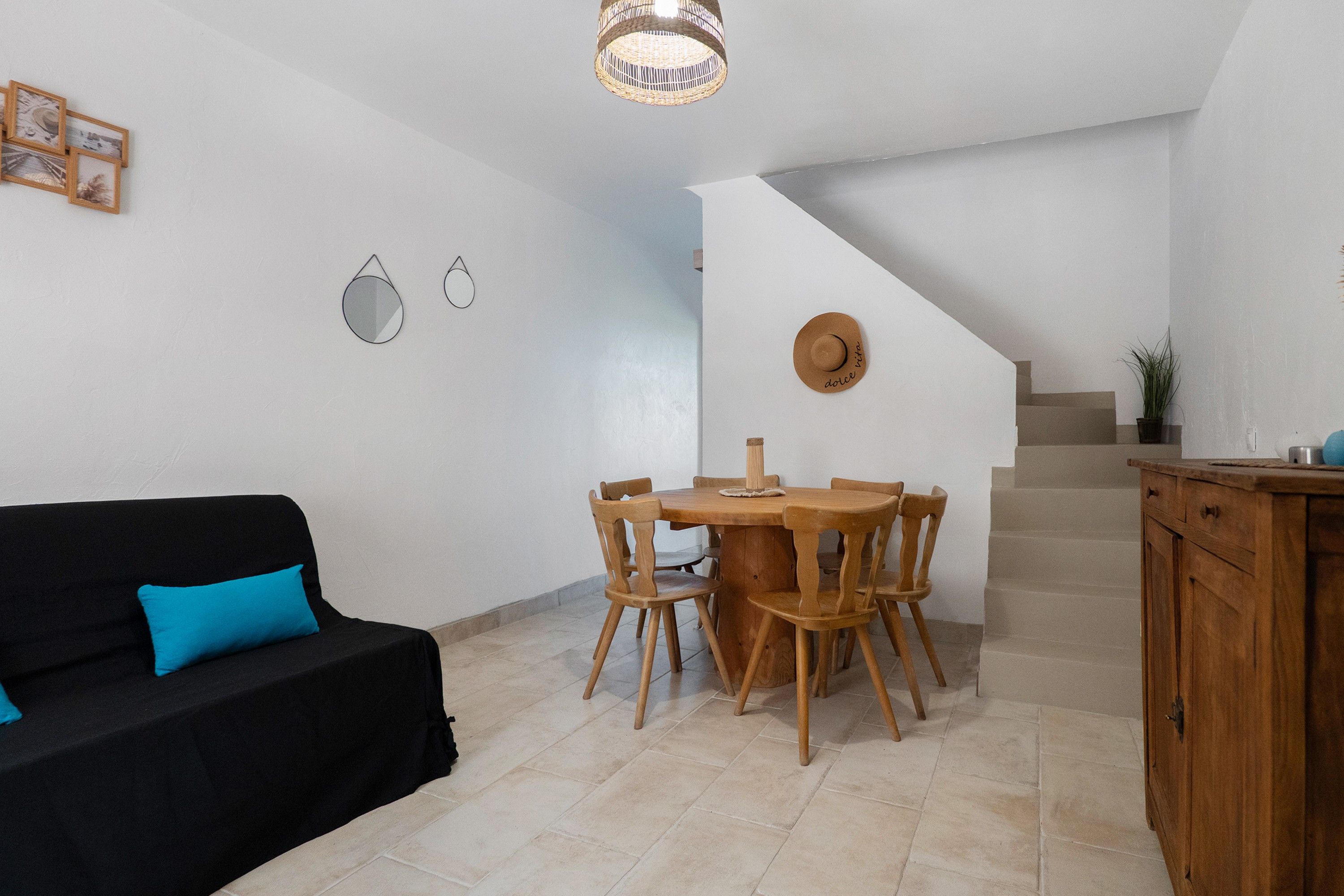 Una sala de estar con un sofá negro, una mesa de comedor de madera y sillas, una escalera y una zona de cocina con armarios.