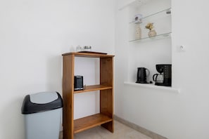 A small kitchenette with a wooden shelf, a trash bin, and a coffee maker on a white wall.