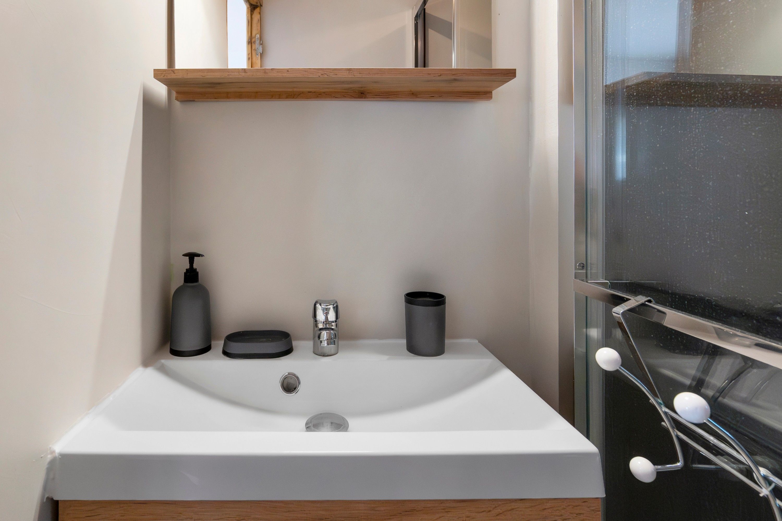 A bathroom with a white sink, a wooden shelf above it, and a glass shower enclosure.