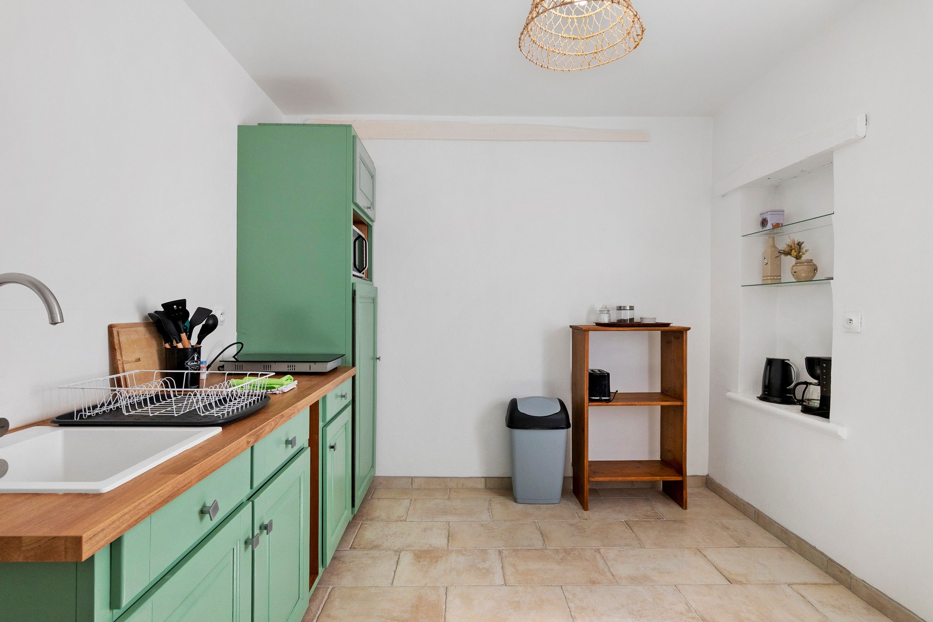 Une cuisine compacte comprenant un évier, des placards verts, un plan de travail en bois, une poubelle et une étagère contenant des ustensiles de cuisine.