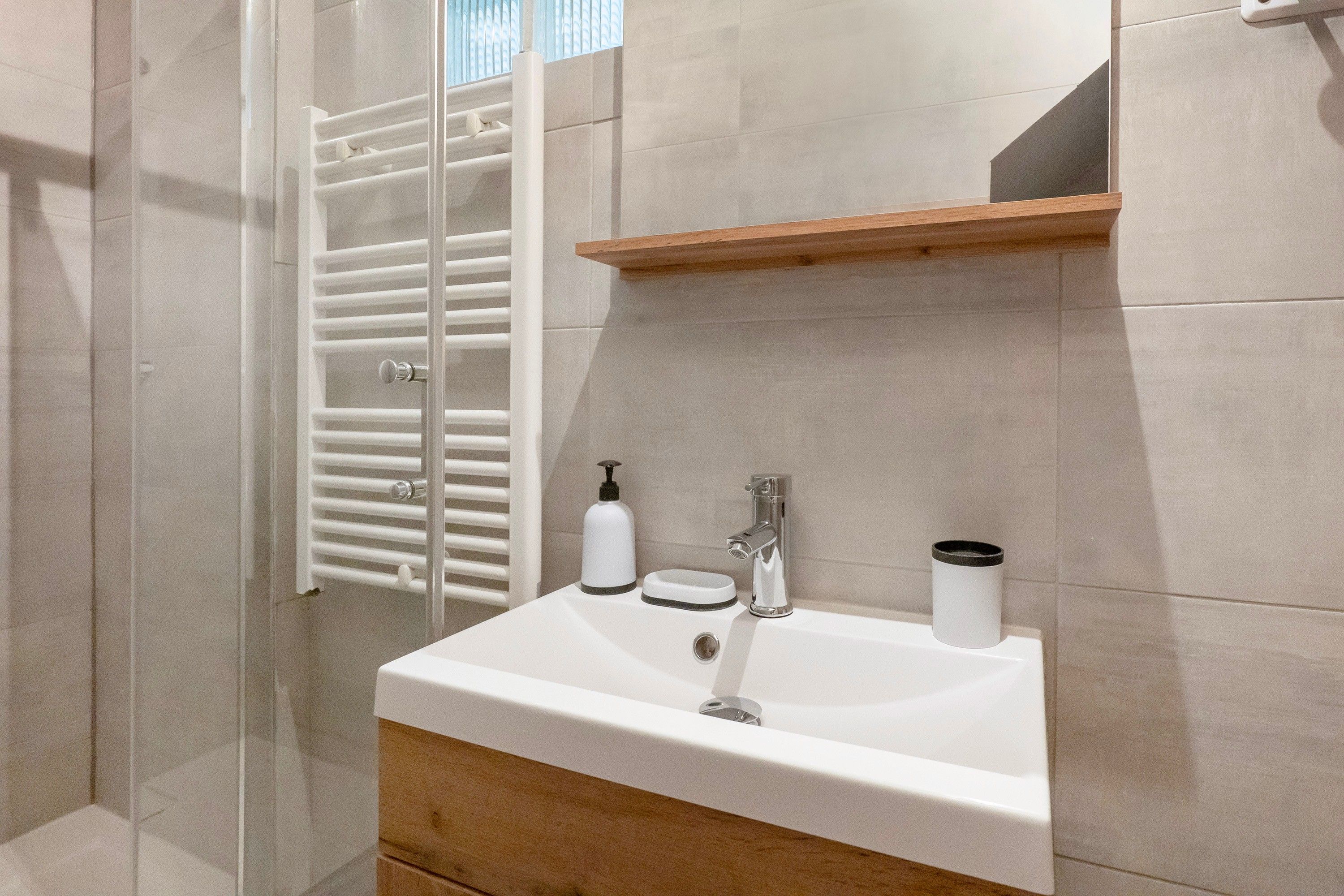 Un baño con lavamanos blanco, un espejo, repisa de madera y cabina de ducha con mampara de vidrio.