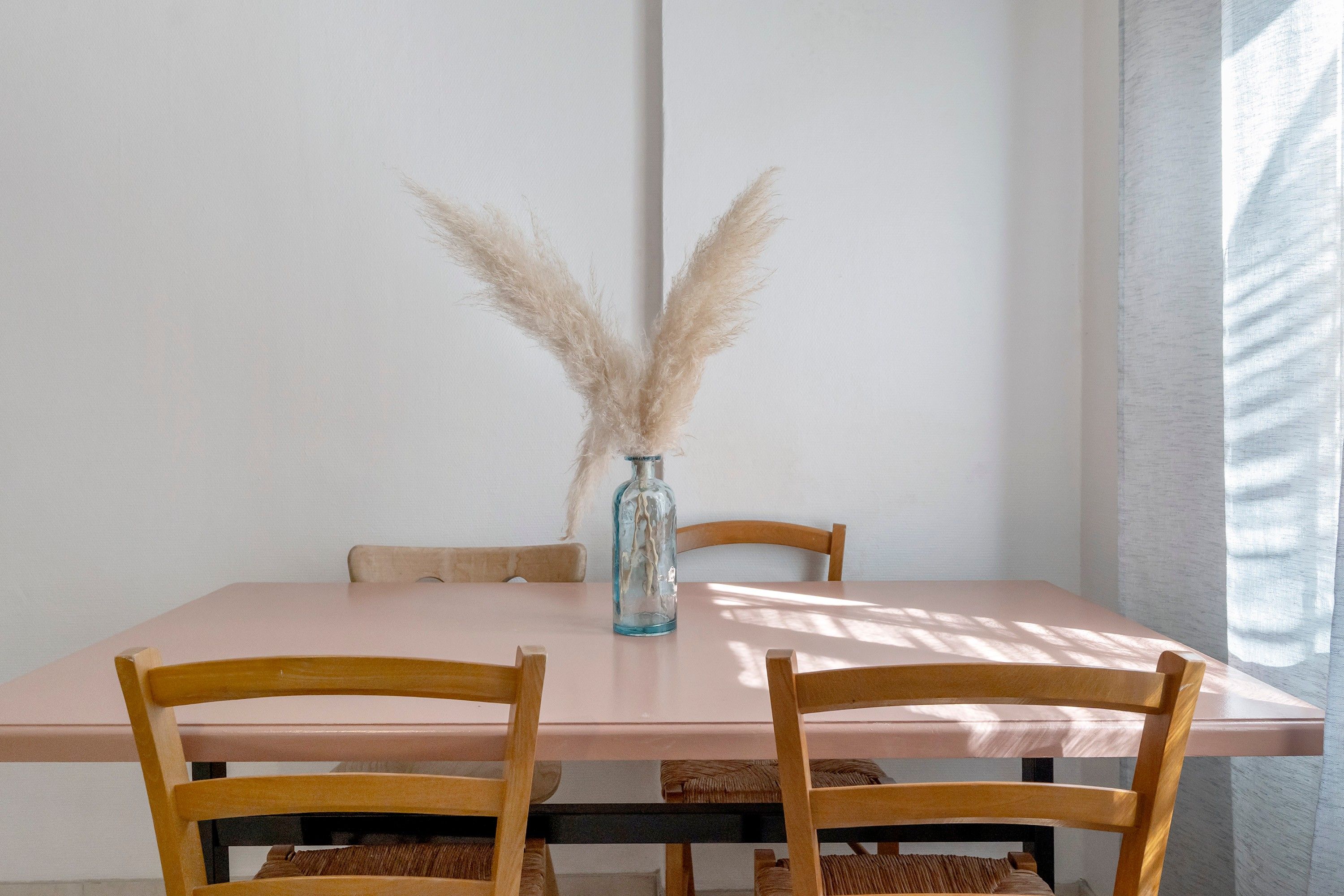 A dining table with a vase of pampas grass, two wooden chairs, and a light-colored tablecloth.