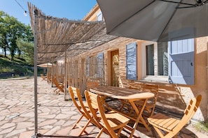 Een terras buiten met houten tafels en stoelen, een rieten dak en een vloer van natuursteen.