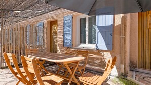 Une terrasse extérieure avec une table et des chaises en bois, un toit de chaume et une fenêtre avec des volets bleus.