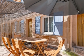 Een terras buiten met een houten tafel en stoelen, een rieten dak en een raam met blauwe luiken.