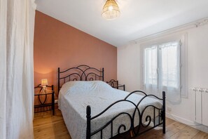 Une chambre à coucher avec un lit, une table de chevet, une lampe, une fenêtre avec des rideaux et un radiateur.