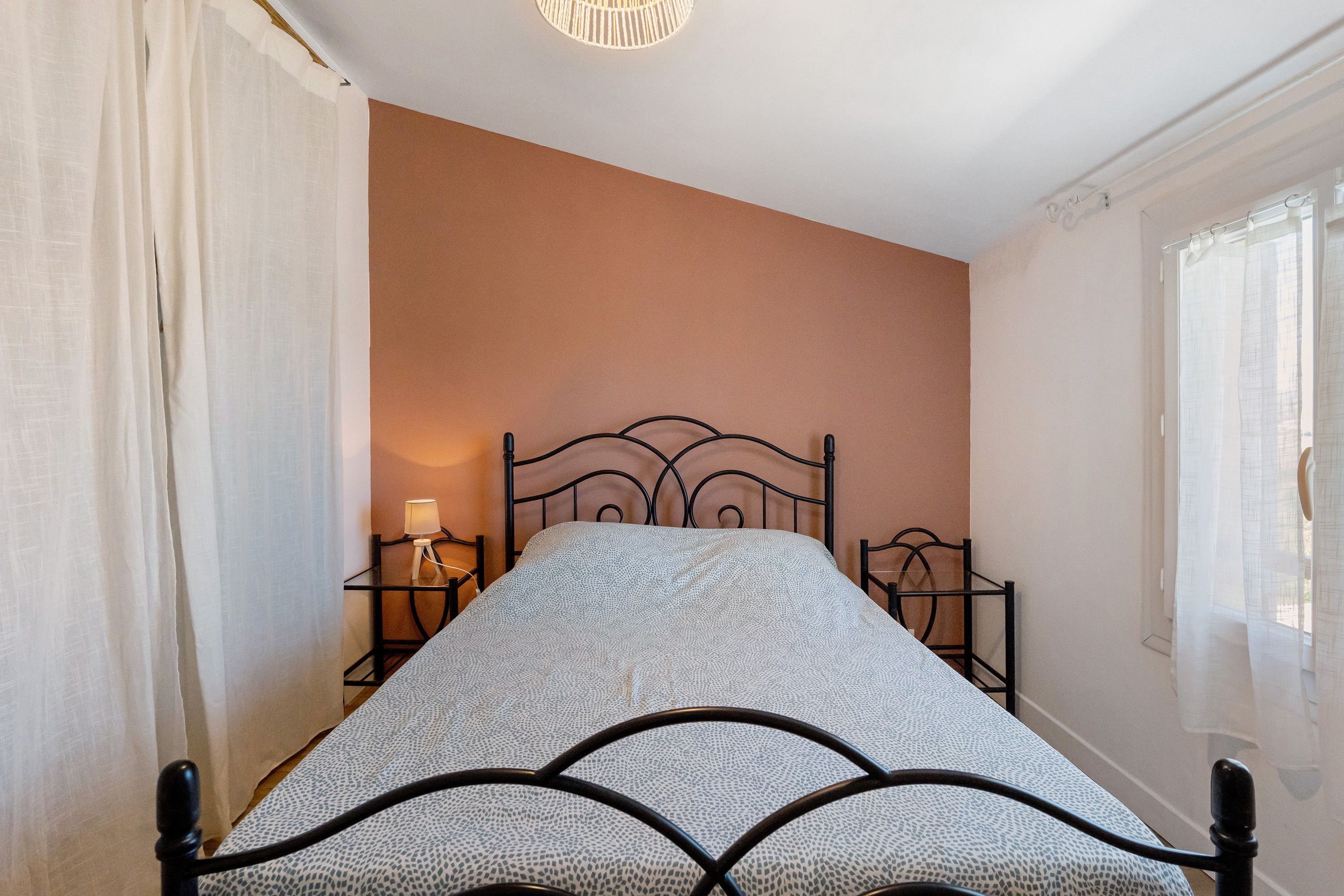 Una cama con estructura metálica y colcha estampada, una mesita de noche con lámpara, y una ventana con cortinas.