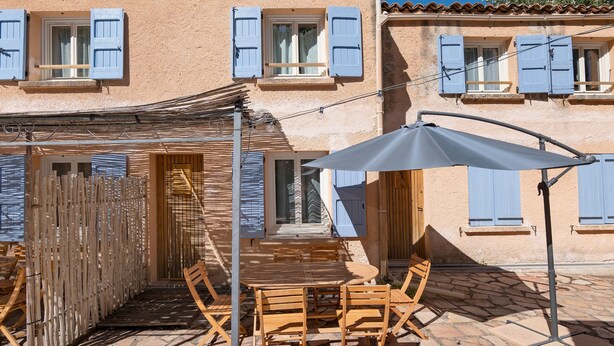 Une terrasse dotée de meubles en bois et d’une table, entourée d’une zone pavée de pierres et d’une clôture en bois.