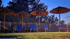 Un espace aménagé au bord de la piscine, avec des chaises longues et des parasols en chaume, entouré d’une clôture en métal et d’arbres.