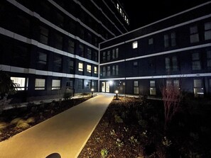 Un immeuble à plusieurs étages, doté d’un chemin central, de fenêtres illuminées et d’un petit espace jardin.