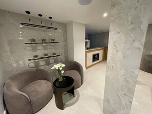 Un espace d’attente moderne avec deux fauteuils gris, une table basse noire ornée d’un vase de fleurs, et un comptoir d’accueil en arrière-plan.