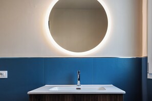 A bathroom with a round mirror, a white sink, and blue walls. - Brezza Apartments (Santa Flavia)