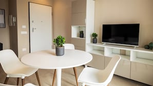 A modern living room with a round white table, two white chairs, a flat-screen TV on a white stand, and a door with a light switch. - Brezza Apartments (Santa Flavia)