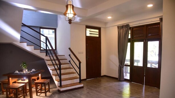 A staircase with wooden steps and black railings, a dining table with chairs, a hanging light fixture, and a large window with curtains. - Belvita Resort Ella (Ella)