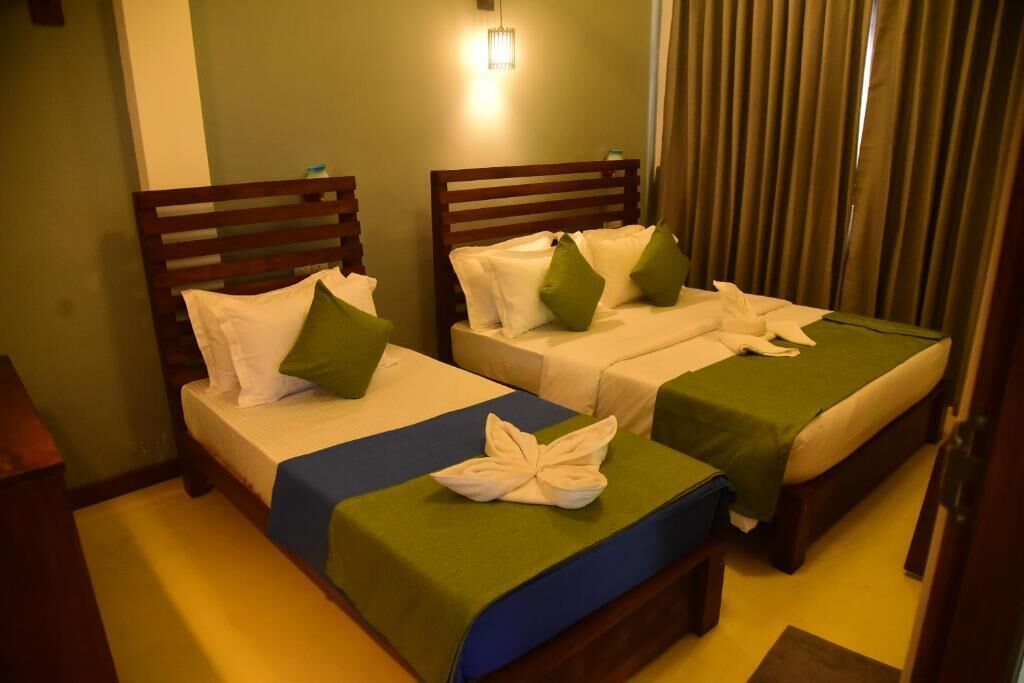 A hotel room with two single beds, each with a wooden headboard, white bedding, and green pillows. There is a small table between the beds, and a light fixture above the headboard.