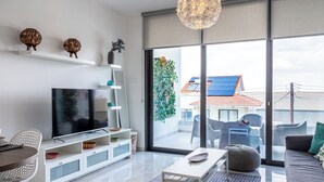 Ein modernes Wohnzimmer mit Flachbildfernseher, weißen Möbeln, einer Glastür, die auf einen Balkon führt, und Blick auf ein Gebäude mit Solaranlage.