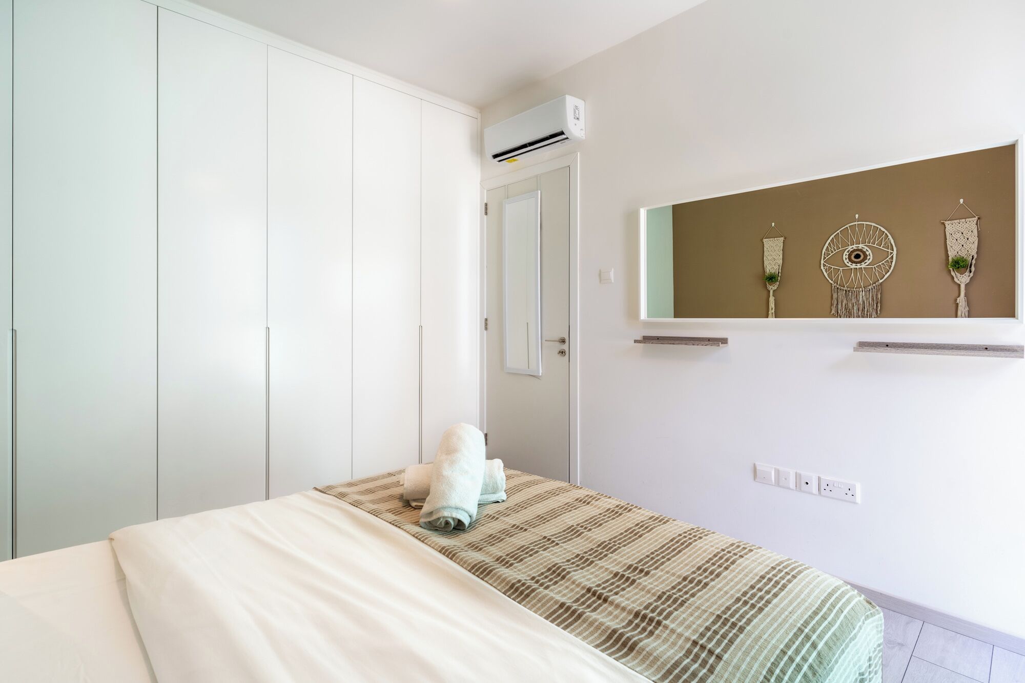 Ein Schlafzimmer mit einem Bett, einem Kleiderschrank, einer Klimaanlage, einem an der Wand befestigten Regal mit Dekoration und einer Tür.