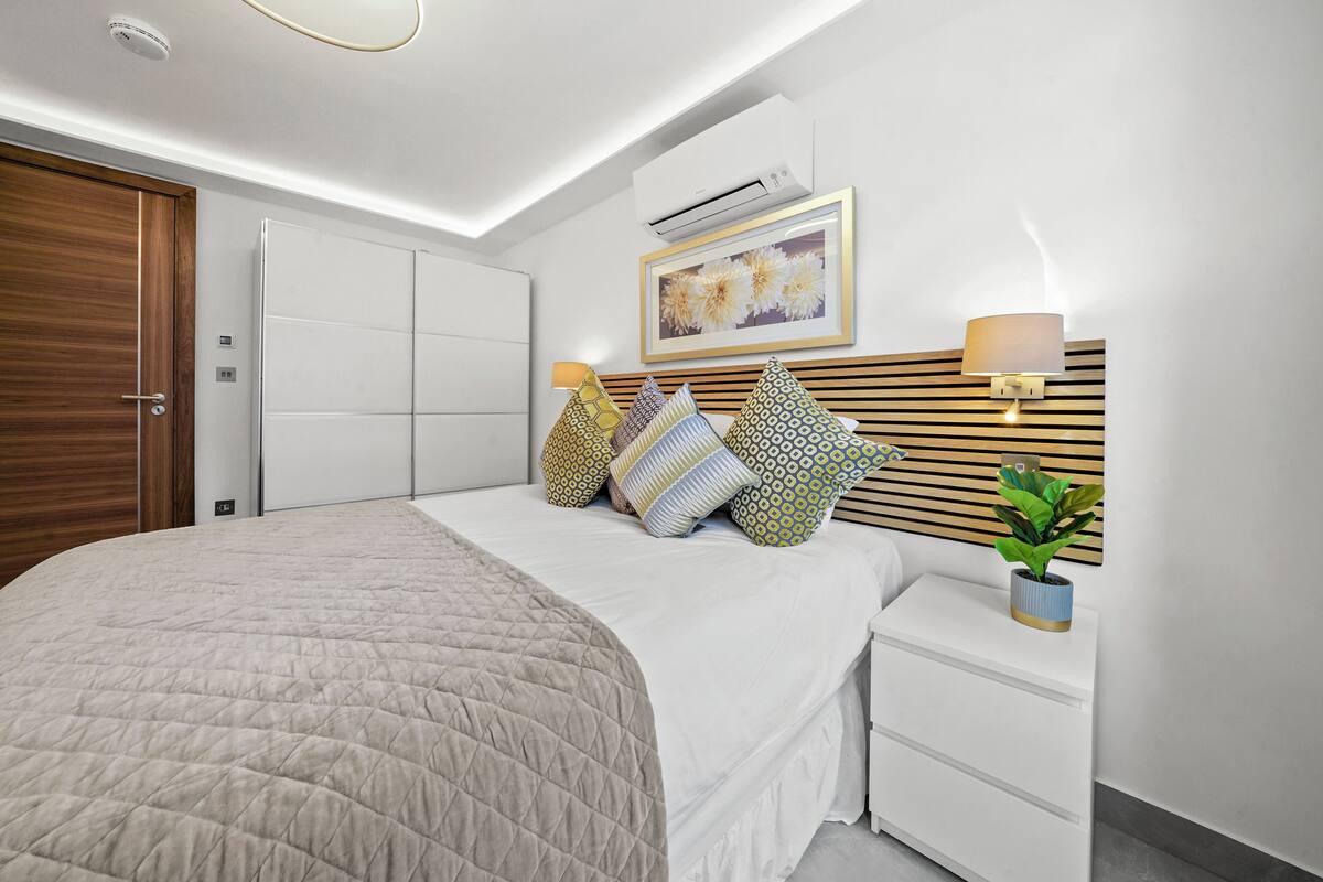 A modern bedroom with a bed, bedside tables, a wall-mounted air conditioner, a framed artwork, and a potted plant.