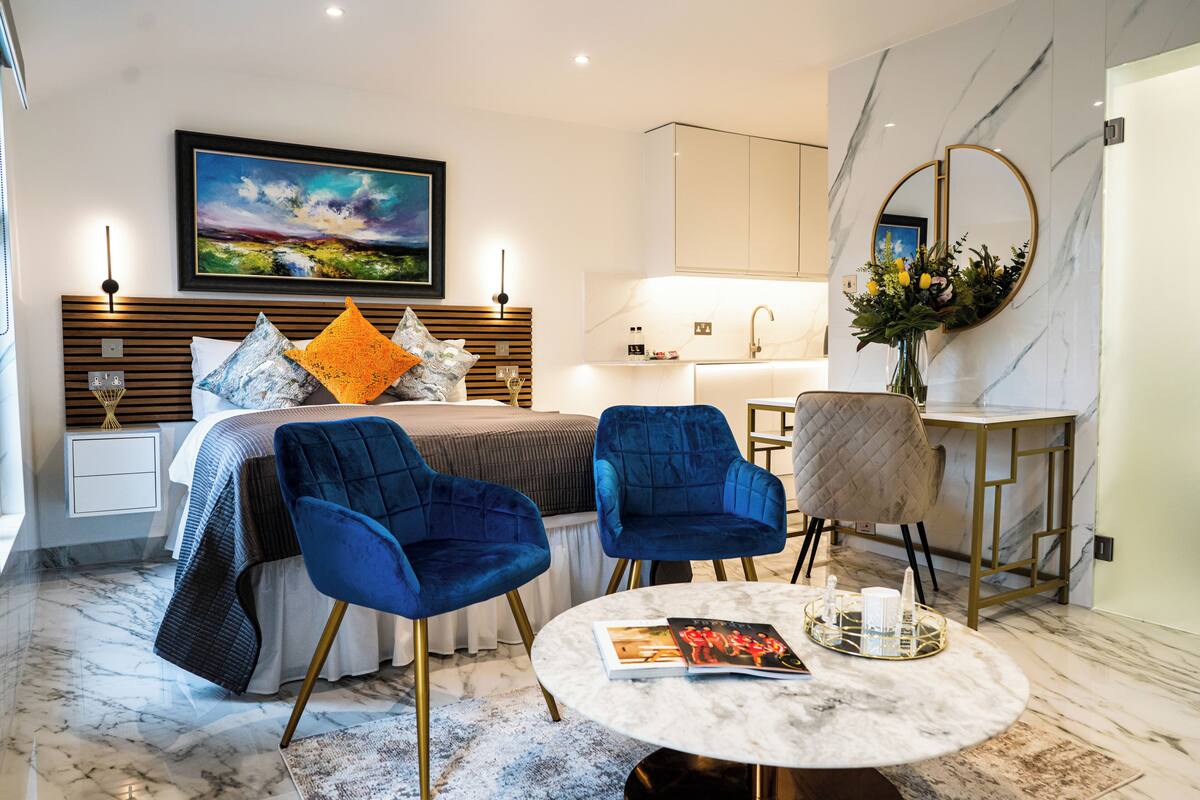 A modern hotel room with a bed, two blue chairs, a round table with a magazine, a painting on the wall, and a kitchen area.