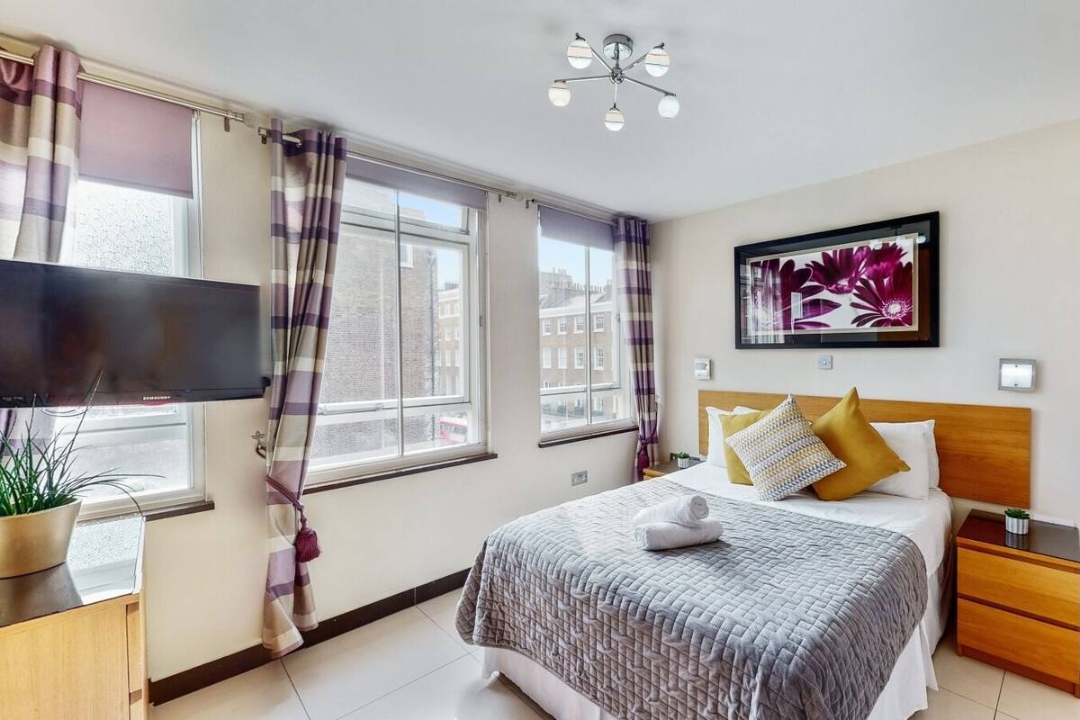 A hotel room with a bed, a TV, a window with curtains, a painting on the wall, and a nightstand.