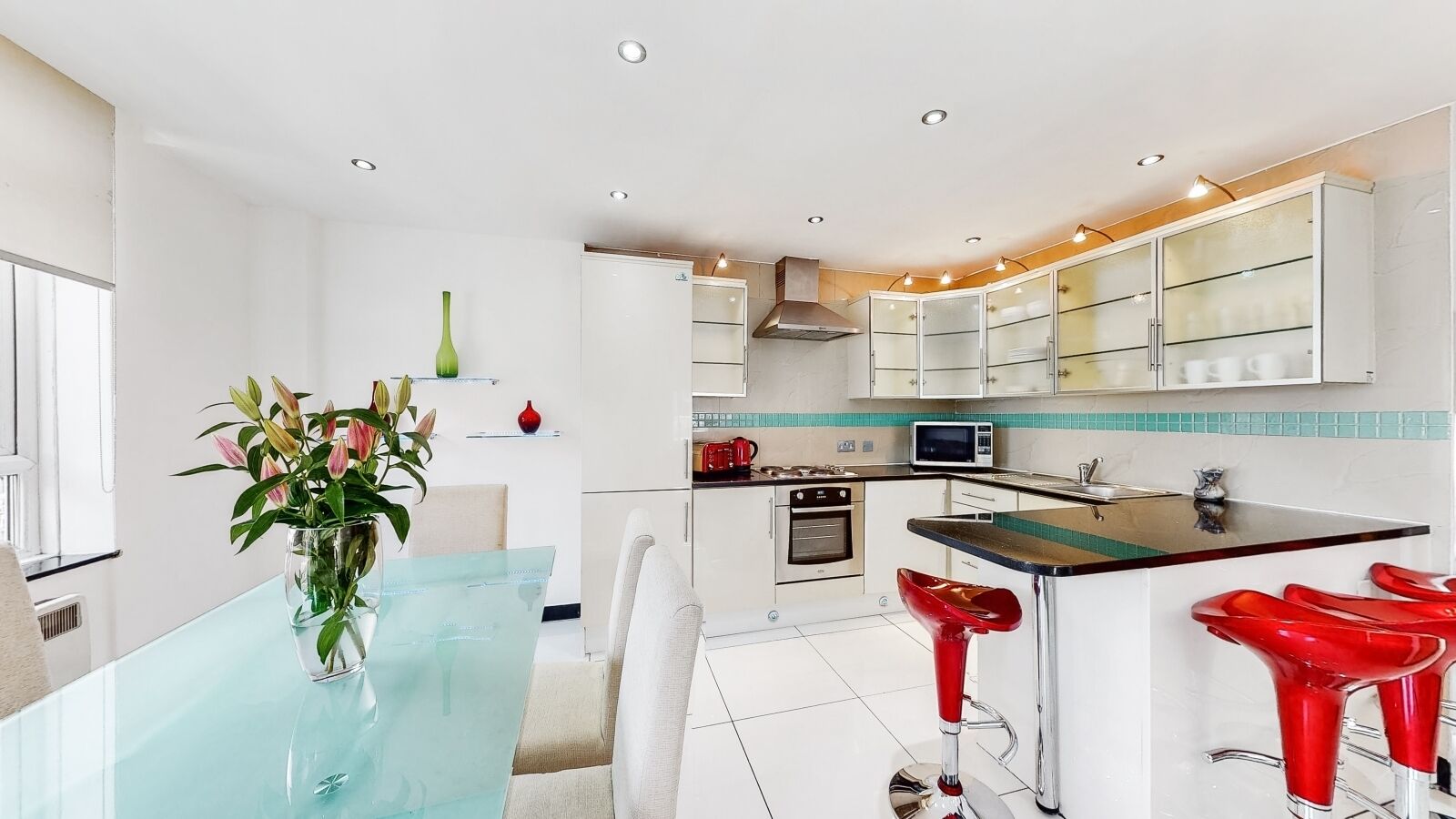 A modern kitchen with white cabinets, a glass dining table, and red bar stools.