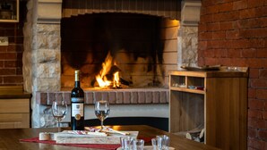 A table with a bottle of wine, glasses, and a wooden board with food items. A brick fireplace with a fire is in the background.