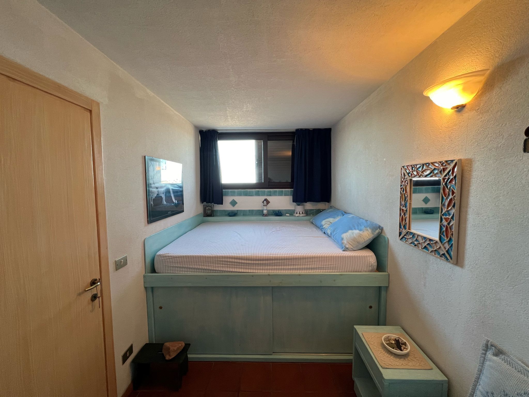 A compact room with a bed, a small table, a window with curtains, a wall-mounted light, and a door.