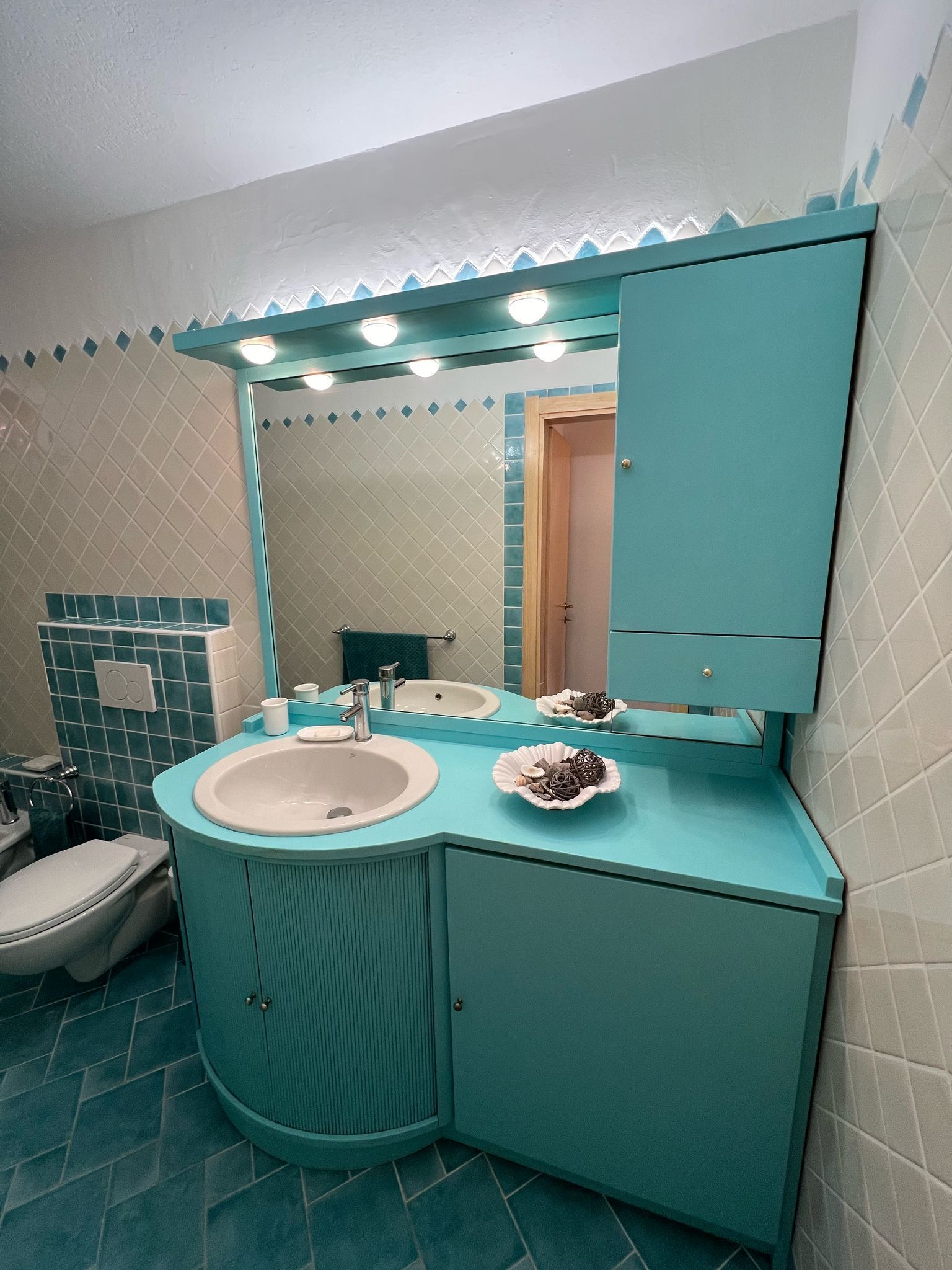 Un baño con tocador turquesa, lavamanos blanco, espejo y retrete.