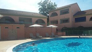 Une piscine entourée de chaises longues et de parasols, le tout bordé d’un bâtiment doté de fenêtres en arc de cercle et d’un toit recouvert de tuiles.