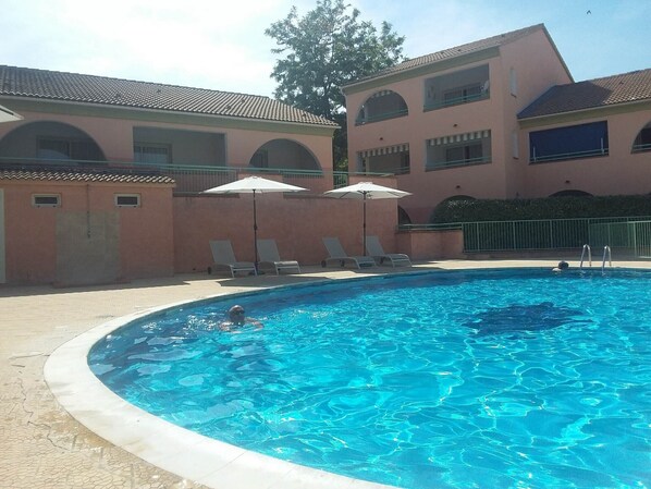 A swimming pool with lounge chairs and umbrellas, surrounded by a building with arched windows and a tiled roof. - Holiday Home 'Apartment Les Jardins De La Mer' with Shared Pool, Wi-Fi and Air Conditioning (Linguizzetta)