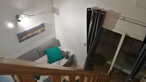 Un escalier mène à un espace de vie comprenant un canapé, un coussin turquoise, un luminaire fixé au mur et une porte vitrée avec des rideaux.