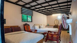 A room with a bed, two chairs, a small table, a fan, and a window with blinds.