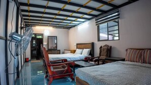A hotel room with two beds, a wooden chair, a small table, a mirror, and a ceiling fan.