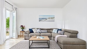 Ein Wohnzimmer mit einem Ecksofa, einem Couchtisch, einem Gemälde von einem Strand und Blick durch eine Glastür.