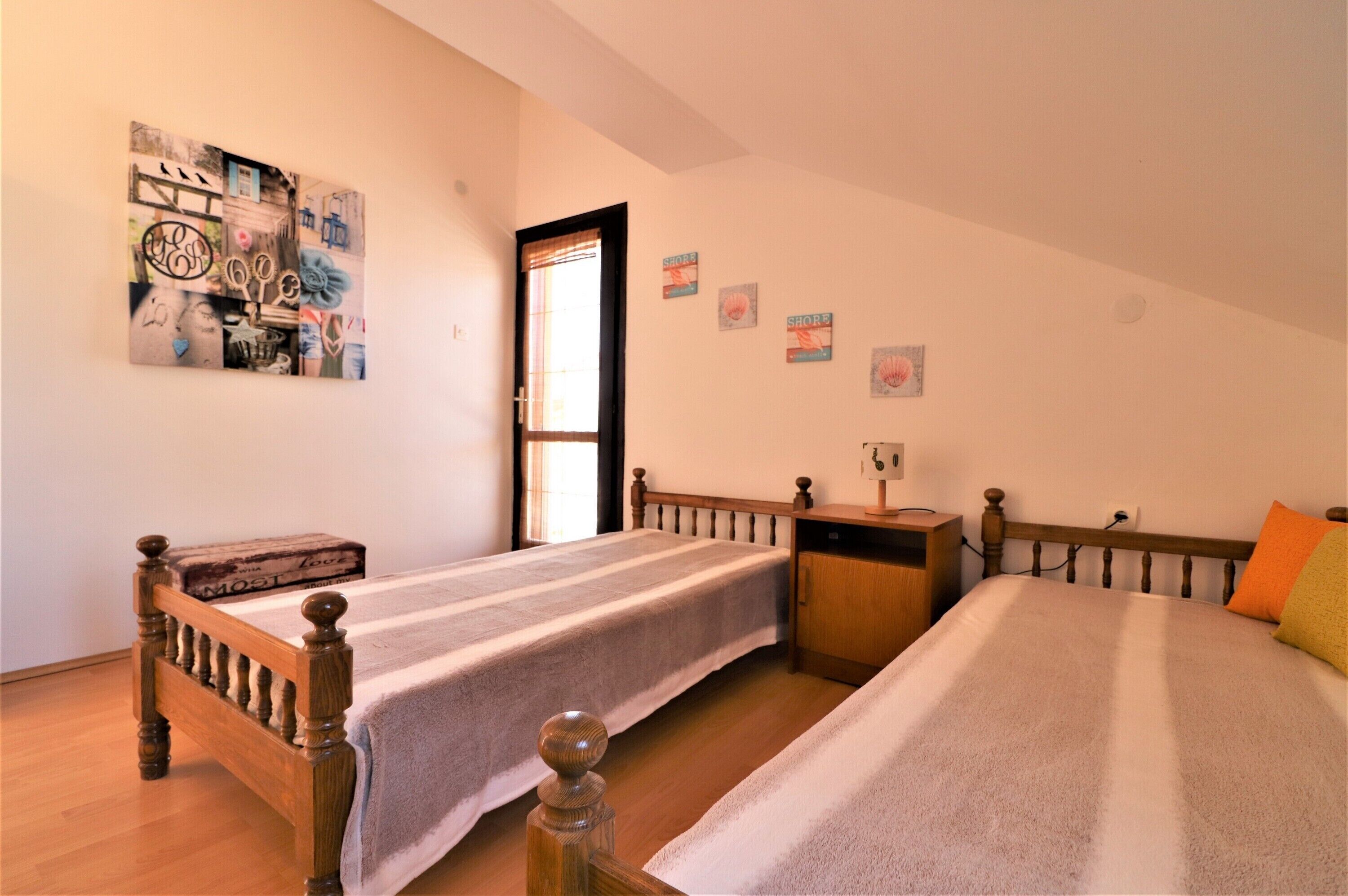 A bedroom with two beds, a wooden headboard, a bedside table, a nightstand, a wall-mounted shelf, and a door with a window.