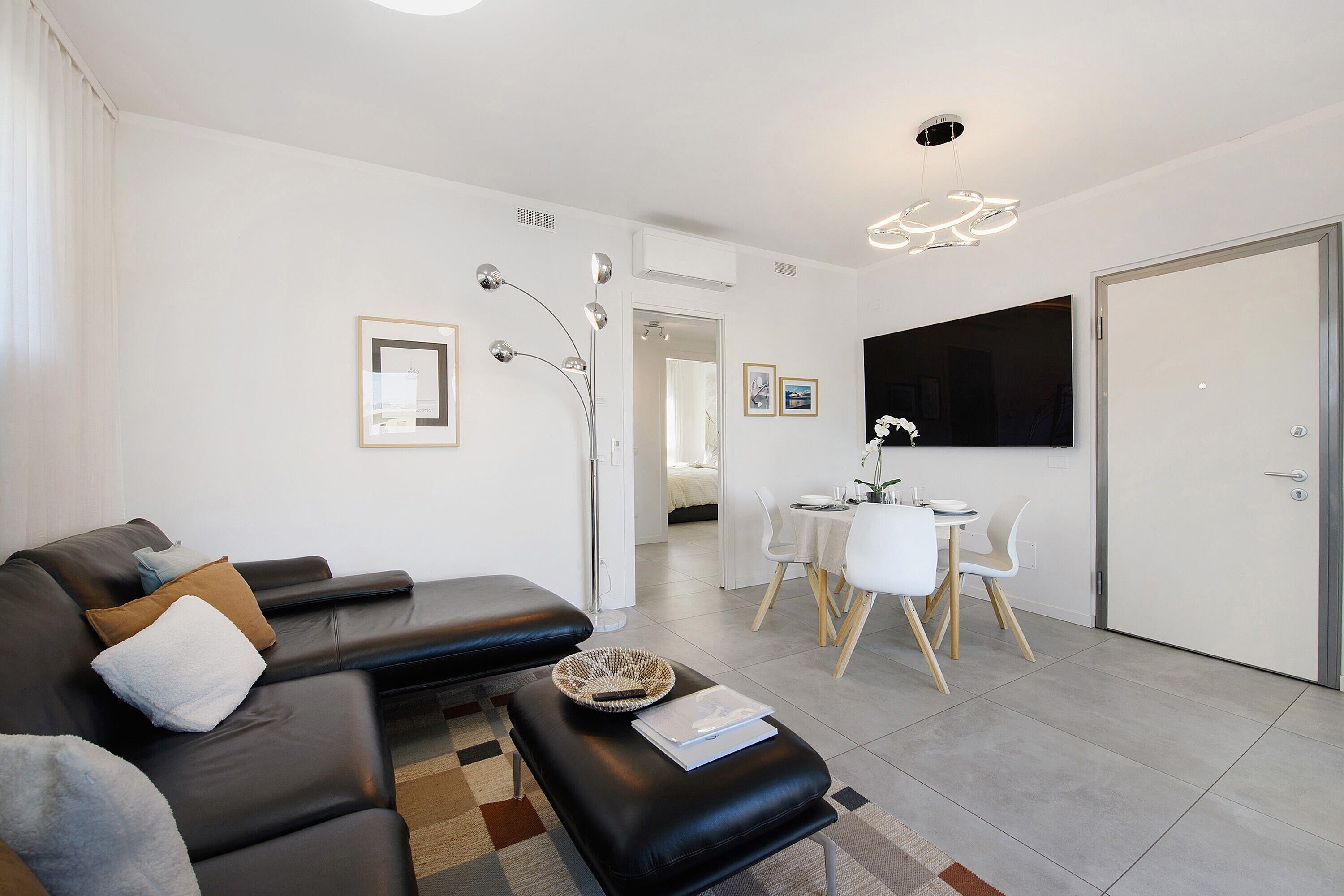 A modern living room with a black leather sofa, a dining area with a white table and chairs, and a large flat-screen TV mounted on the wall.