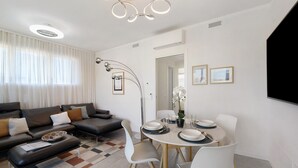 Una sala de estar moderna con sofá, mesa de comedor y sillas, lámpara de techo y un amplio ventanal con cortinas.