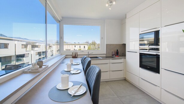 Una cocina moderna con comedor junto a un ventanal grande, que incluye un horno empotrado, un lavabo y una mesa de comedor preparada con platos y cubiertos.