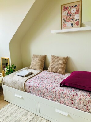 A bedroom with a sloped ceiling, a bed with floral bedding, two textured pillows, a red accent pillow, a white bedside table, and a framed floral artwork on the wall.