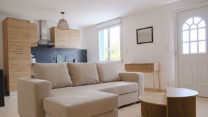Un salon moderne avec un canapé, une petite table ronde et un coin cuisine doté de meubles en bois et d’un luminaire suspendu.