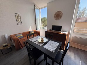 A cozy living room with a sofa, a dining table with chairs, a TV on a cabinet, and a window with a view. - 250-metre walk from the spa, superb bright T1bis 3-person apartment (Eugénie-les-Bains)