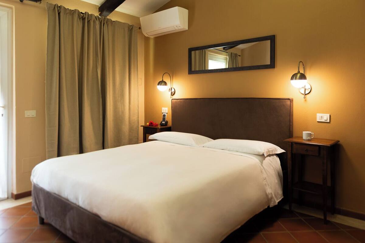 A hotel room with a large bed, two bedside lamps, a nightstand with a cup, a mirror above the bed, and an air conditioning unit.