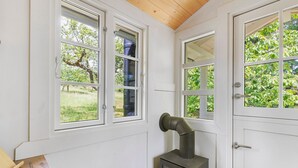 Une pièce aux murs blancs, avec un plafond en bois et un poêle à bois. Elle comporte trois fenêtres à cadre blanc, donnant sur un paysage verdoyant.